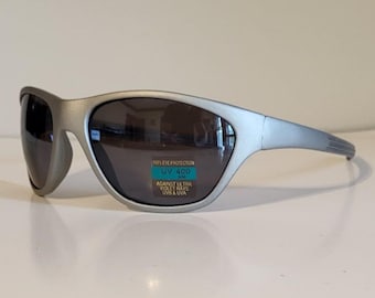 Może przedstawiać: Srebrne okulary przeciwsłoneczne z ciemnymi soczewkami. Okulary mają zakrzywioną konstrukcję i małą naklejkę z napisem "100% EYE PROTECTION UV 400 AGAINST ULTRA VIOLET RAYS UVB & UVA".