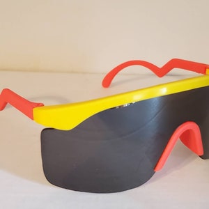 Peut inclure: Lunettes de soleil en plastique jaune et rouge avec un design futuriste. Les verres sont gris foncé et la monture a une forme ondulée.