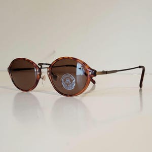 Peut inclure: Lunettes de soleil ovales avec une monture à motif écaille de tortue et des verres marron. Les lunettes ont un pont et des branches en métal. Les verres ont un autocollant qui dit "ULTIMATE 400 LENSES SPECIAL PURPOSE".