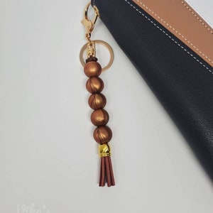 May include: A gold key chain with a brown tassel and a string of copper-colored beads. The key chain is attached to a black, brown, and white leather wallet.