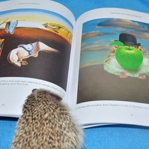 May include: An open book with two pages showing different paintings. The left page shows a painting of a melting clock and a hedgehog. The right page shows a painting of a hamster wearing a hat and holding an apple. The book is on a blue surface.