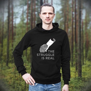 May include: Black hoodie with a white graphic of a paintbrush and the text "THE STRUGGLE IS REAL". The hoodie has a hood and a front pocket. The person is wearing blue jeans.