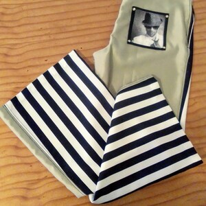 May include: A pair of green trousers with a black and white striped fabric on the bottom. The trousers have a patch pocket with a photo of a person wearing a hat and sunglasses.