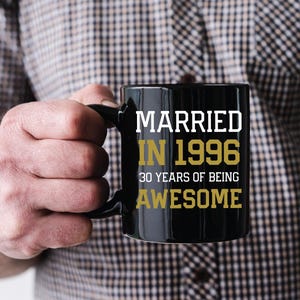 May include: Black coffee mug with the text "MARRIED IN 1996 30 YEARS OF BEING AWESOME" in white and gold lettering. The mug is held by a person wearing a plaid shirt.