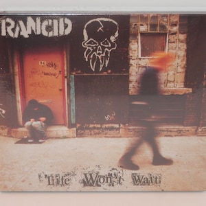 May include: A CD cover for the album "Life Won't Wait" by the band Rancid. The cover features a grainy photo of a brick wall with a door and a skull graffiti. A person walks in the foreground, and another person sits on the steps.