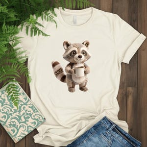 May include: Cream-colored t-shirt featuring a cartoon raccoon holding a coffee cup. The raccoon has a brown and gray color scheme with a patterned tail. The shirt is paired with blue denim jeans.