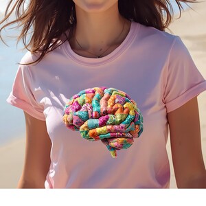 May include: A light pink t-shirt featuring a colorful, patchwork-style brain graphic. The brain design incorporates various fabric patterns and colors, including pink, blue, green, and yellow. The shirt has short sleeves and a crew neck.