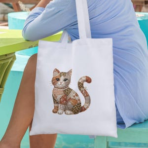 May include: White tote bag featuring a patchwork cat design. The cat has a mix of patterns, including leopard print and plaid, with a brown and orange color scheme. The bag is carried by a person wearing a light blue shirt.