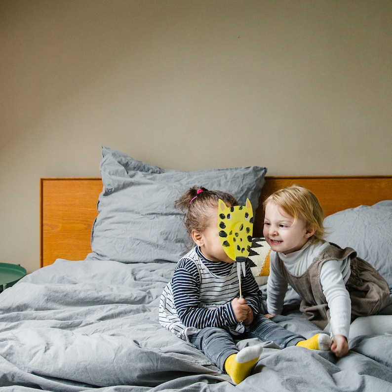 May include: Two children are playing on a bed with a yellow and black animal mask. The child on the left is wearing a striped shirt and the child on the right is wearing a brown dress.