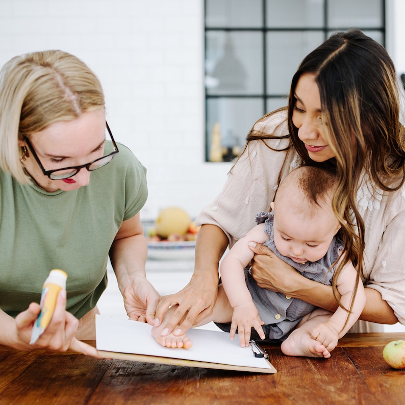 May include: Two people, one holding a baby, are making a footprint on a white piece of paper. The baby is wearing a gray outfit and is being held by a woman with long brown hair. The woman is wearing a light beige top. The other person is holding a yellow and blue tube of paint.