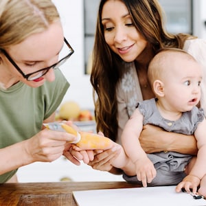 May include: A woman is feeding a baby pureed food from a pouch. The baby is sitting on the lap of another woman. The baby is looking at the camera with a curious expression.
