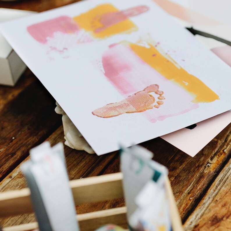 May include: A white card with pink and yellow paint handprints and footprints. The card is on a wooden surface.