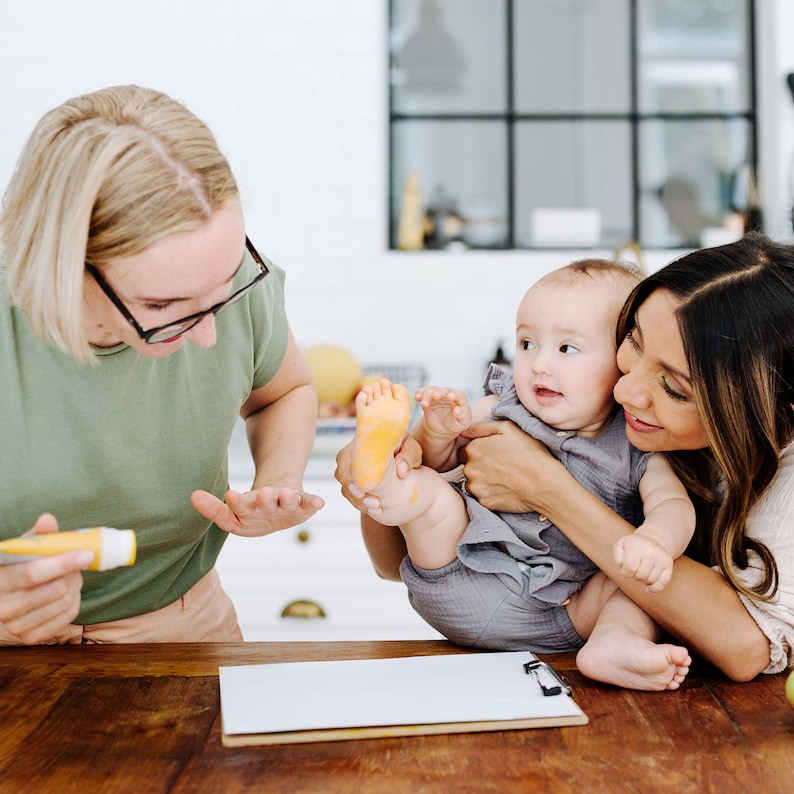 May include: Two adults are making handprints with a baby. The baby is wearing a gray outfit and has a yellow handprint on their foot. The adults are holding the baby's hands and feet. There is a white piece of paper on a wooden table.