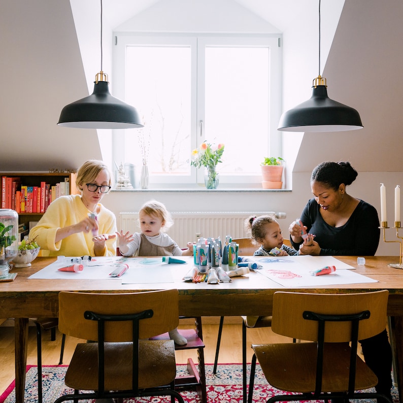 May include: A group of people are gathered around a table, working on art projects. There are paint tubes, brushes, and paper on the table. The people are all smiling and having fun.