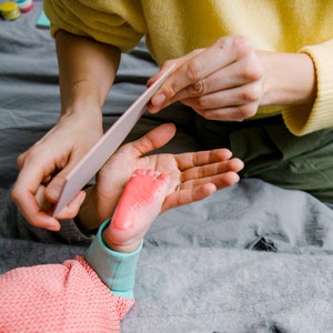 May include: A person holding a baby's foot with pink paint on it. The person is using a white tool to smooth out the paint.