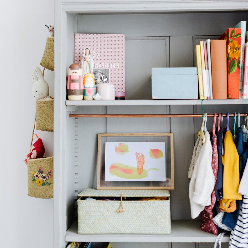 May include: A white bookshelf with a copper rod for hanging clothes. The shelf is filled with books, a framed print of a child's footprint, a wooden toy, and a wicker basket. The print is in shades of green, yellow, and pink.