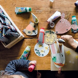 May include: A table with various craft supplies, including paint tubes, paintbrushes, and paper cutouts. The cutouts include a fox, a deer, a bunny, and a beehive. The table is made of wood and has a light brown finish.