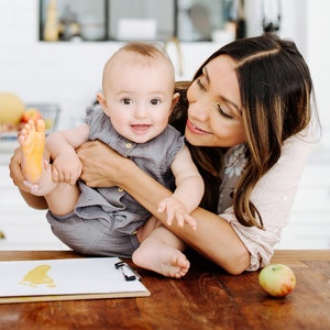 May include: A woman holds a baby in her arms, smiling at the camera. The baby has a yellow footprint on its foot and is looking at the camera. A piece of paper with a yellow footprint on it is on the table in front of them, along with a green apple.