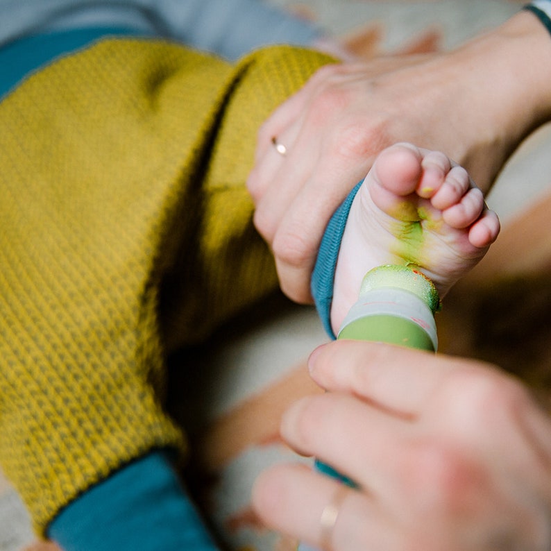 May include: A person's hand holding a green paint roller and applying green paint to a baby's foot. The baby is wearing a yellow and blue striped shirt.