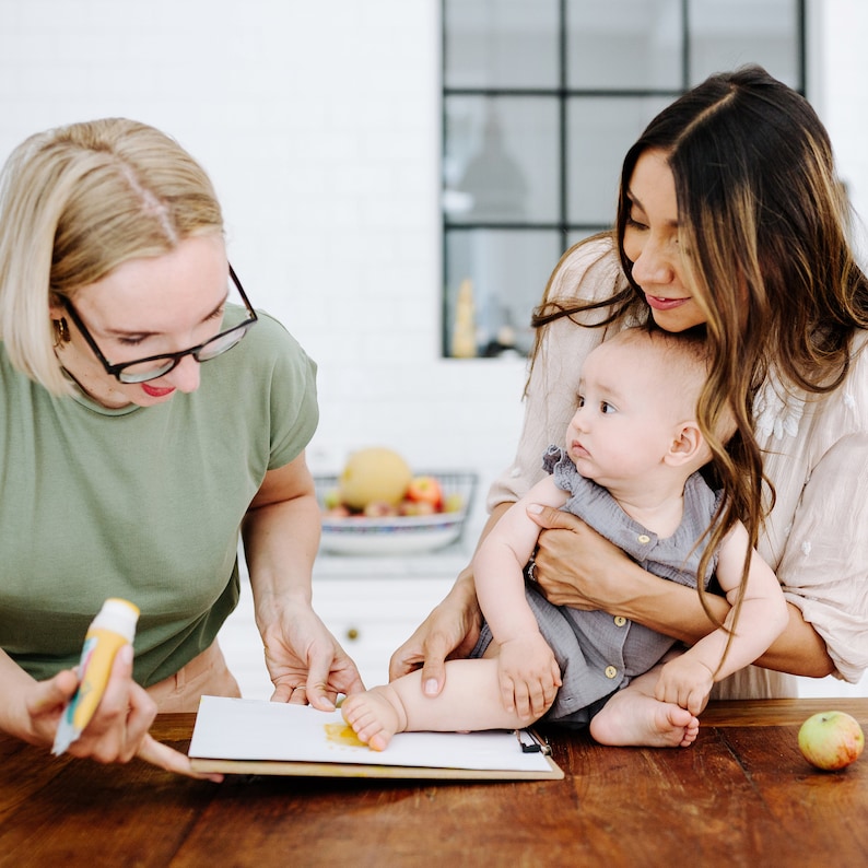 May include: A woman holds a baby while another woman uses a tube of paint to make a handprint on a piece of paper. The baby is wearing a gray onesie and the woman holding the baby is wearing a white shirt. The woman making the handprint is wearing a green shirt and glasses.