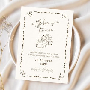 May include: A white baby shower invitation with a brown border and a drawing of two cinnamon rolls. The text reads "a little bun is in the oven" and includes the date and time of the shower, "01.30.2030 @2PM", and the location, "10 Anywhere St. Any City".
