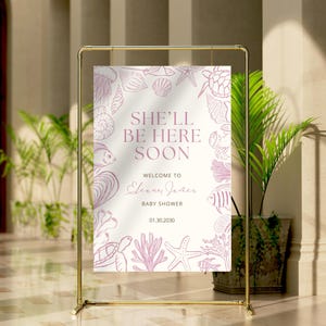 May include: A baby shower sign with a gold frame. The sign has a cream background with pink seashell and sea creature illustrations. Text reads "She'll Be Here Soon" and "Welcome to Elena Jones Baby Shower 01.30.2030".