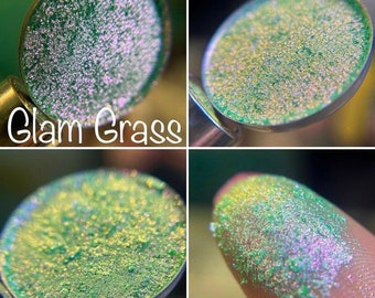 May include: A close-up of a green and gold iridescent eyeshadow with a shimmery finish. The eyeshadow is in a small round container and is labeled "Glam Grass".