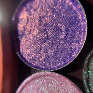 May include: Two round, shimmery eyeshadows. The top eyeshadow is a deep purple with a fine, iridescent glitter. The bottom eyeshadow is a light pink with a fine, iridescent glitter.
