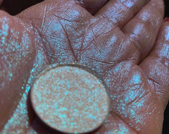 May include: A white iridescent glitter eyeshadow with a blue and green shimmer. The glitter is loose and scattered on a hand.