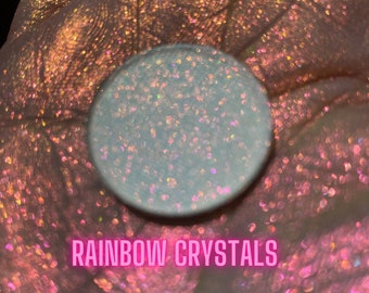 May include: A close-up of iridescent rainbow glitter crystals in a white circle on a hand. The text "RAINBOW CRYSTALS" is written in pink below the circle.