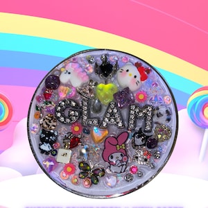 Puede incluir: Un compacto redondo transparente con un borde plateado y un fondo blanco. El compacto está lleno de purpurina de colores, pedrería y pequeños amuletos, incluyendo un unicornio, un personaje de Hello Kitty, una mariposa y una flor. La palabra "GLAM" está escrita en letras plateadas con pedrería. El compacto está etiquetado como "CUSHION FOUNDATION "JUNK CASE"".