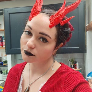 May include: A set of bright red, textured, pointed horns attached to a thin black headband. The horns feature a crinkled, organic surface texture with multiple upward-curving points, designed to be worn on the head as a decorative accessory.