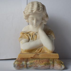 Puede incluir: Busto color crema de una mujer con cabello rizado, apoyando la barbilla en su mano. Viste un vestido amarillo y está sentada sobre un libro con patrones florales decorativos. La escultura es un objeto decorativo.