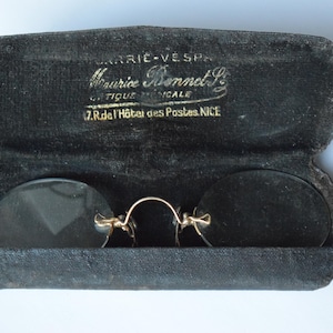 Könnte beinhalten: Vintage runde Brille mit goldfarbenem Steg und dunklen Gläsern, präsentiert in einem offenen, schwarzen Samtetui. Das Etui trägt goldfarbene Schrift: "Maurice Bonnet Optique Nice".