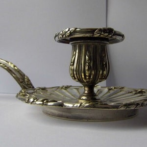 May include: A silver candlestick with a decorative handle and a scalloped base. The candlestick has a fluted design and a floral motif.