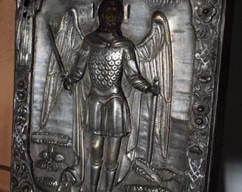 May include: A silver-toned religious icon depicting a figure with wings, holding a sword. The figure is clad in armor and stands within a decorative frame with floral and geometric patterns. The icon is mounted on a wooden easel.