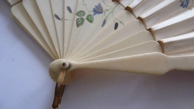 May include: A white folding fan with a floral design. The fan is closed and the handle is made of gold metal.