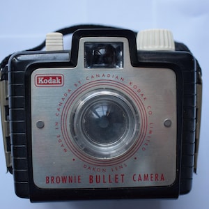 May include: Vintage Kodak Brownie Bullet Camera. The camera has a black body with a silver faceplate. The faceplate has red lettering that reads "Made in Canada by Canadian Kodak Co. Limited" and "Brownie Bullet Camera". The camera has a black strap.