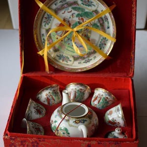May include: A vintage tea set in a red box. The set includes a teapot, six cups, and a plate, all with a detailed, colorful design. The box is lined with red fabric and tied with a yellow ribbon.