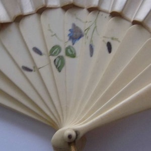 May include: A white folding fan with a floral design. The fan has a blue flower with green leaves and a gold handle.