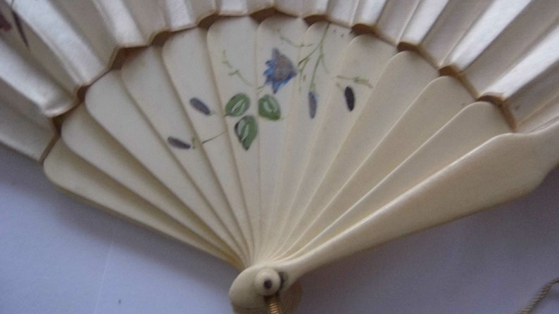 May include: A white folding fan with a floral design. The fan has a blue flower with green leaves and a gold handle.