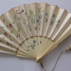 May include: A white hand fan with a floral design. The fan is made of paper and has a wooden handle. The fan has a tassel attached to the handle.