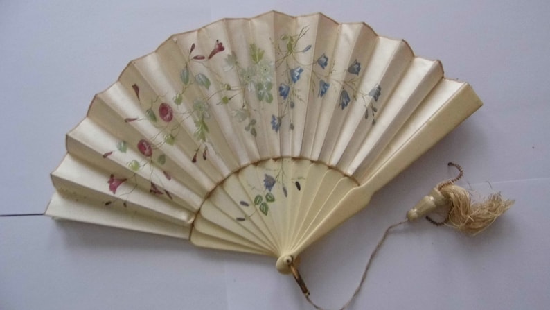 May include: A white hand fan with a floral design. The fan is made of paper and has a wooden handle. The fan has a tassel attached to the handle.