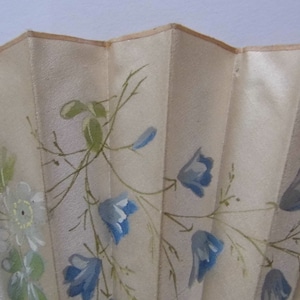 May include: A white silk hand fan with a delicate floral design. The fan features blue and green flowers painted on a white background.