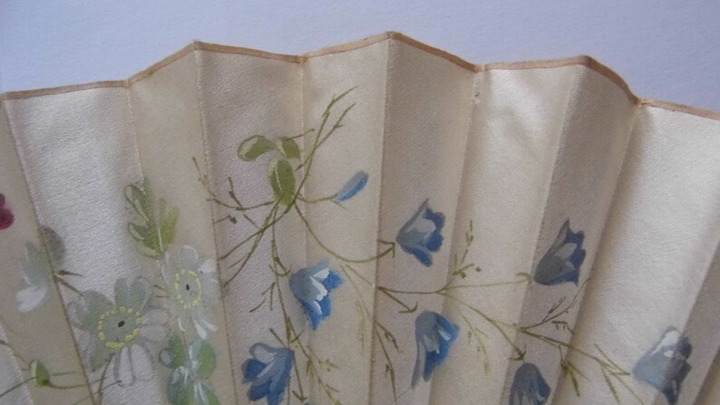 May include: A white silk hand fan with a delicate floral design. The fan features blue and green flowers painted on a white background.
