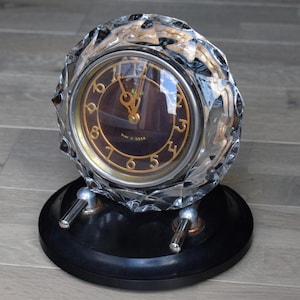 Peut inclure: Une horloge vintage avec un boîtier en cristal facetté et un cadran marron foncé avec des chiffres dorés. L'horloge repose sur une base noire circulaire avec des supports argentés. Le cadran indique "Made in USSR".