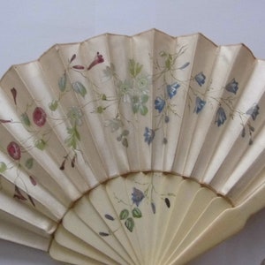 May include: A white hand fan with a delicate floral design painted in shades of pink, blue, and green. The fan is closed and has a tassel attached to the handle.