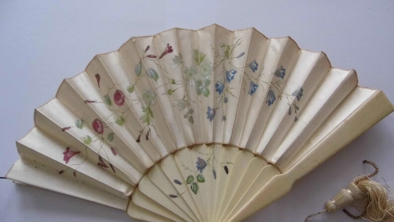 May include: A white hand fan with a delicate floral design painted in shades of pink, blue, and green. The fan is closed and has a tassel attached to the handle.