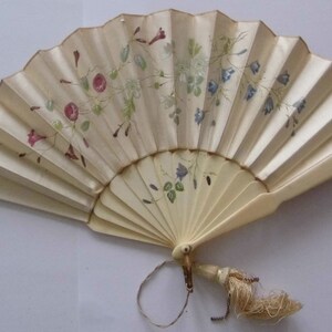 May include: A white folding fan with a floral design in shades of pink, blue, and green. The fan has a tassel attached to the handle.