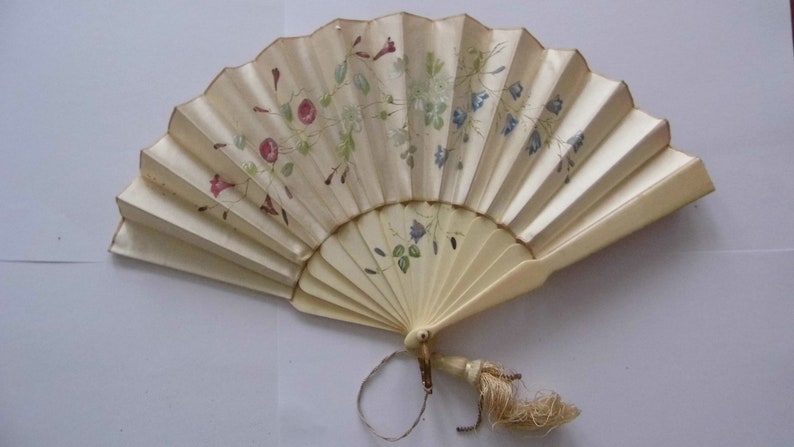 May include: A white folding fan with a floral design in shades of pink, blue, and green. The fan has a tassel attached to the handle.